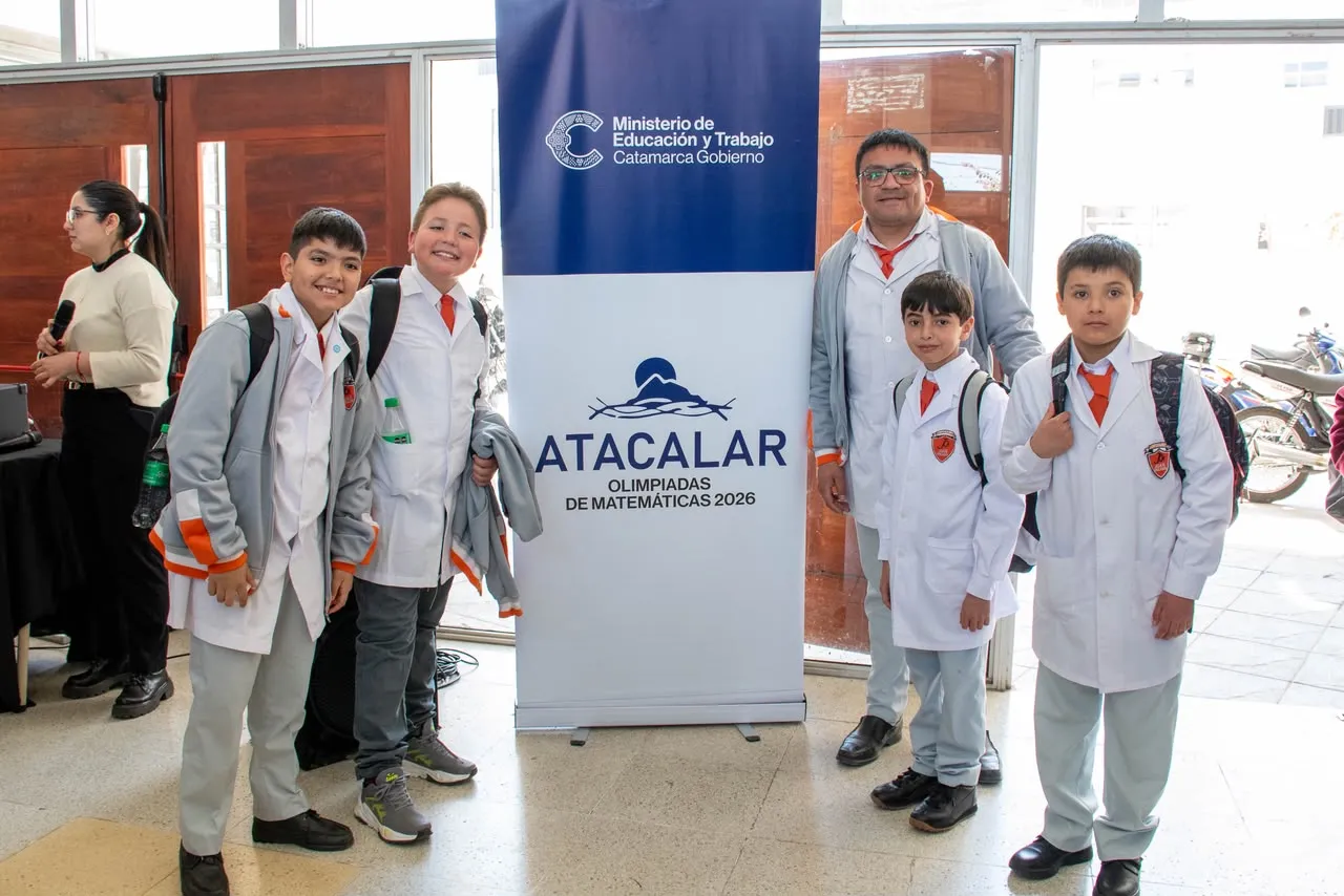 Cuatro chicos y un adulto con guardapolvos blancos posan junto a un banner de olimpíadas de matemáticas.