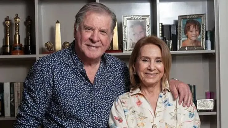 Un hombre y una mujer sonríen abrazados delante de una biblioteca con portarretratos y estatuillas.