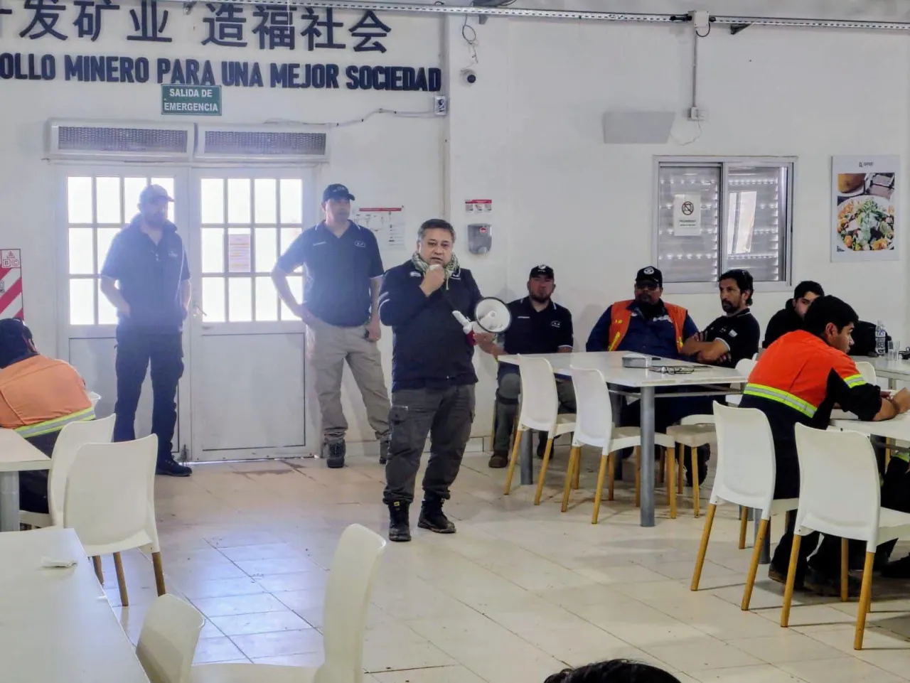 Un hombre habla por megáfono rodeado por varios operarios sentados y parados en un comedor con carteles mineros.
