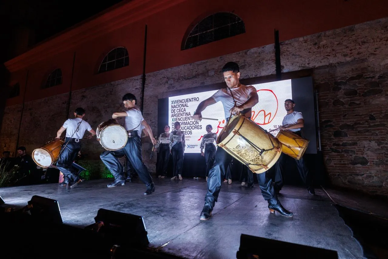 Bailarines folclóricos tocan bombos en un escenario frente a una pantalla.