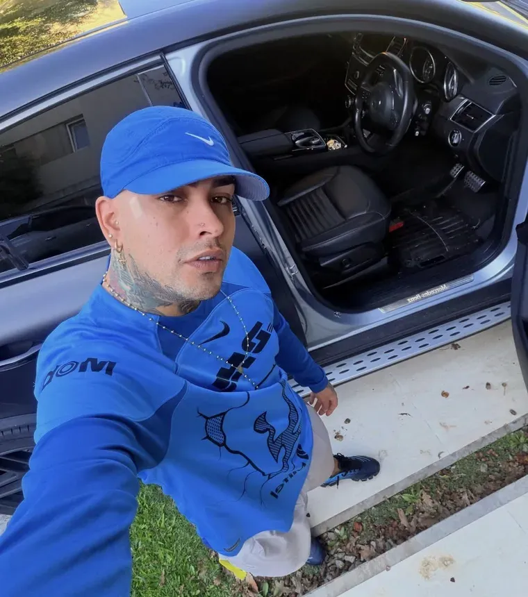 Un hombre con gorra azul, remera manga larga y tatuajes posa parado junto a la puerta abierta de un auto.