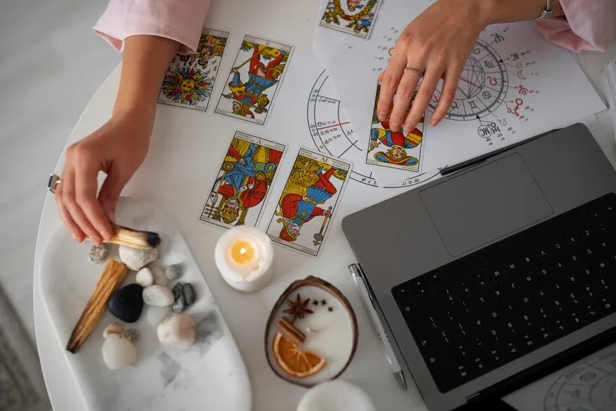 Manos sobre una mesa blanca con cartas de tarot, una carta astral, una notebook, velas y piedras.