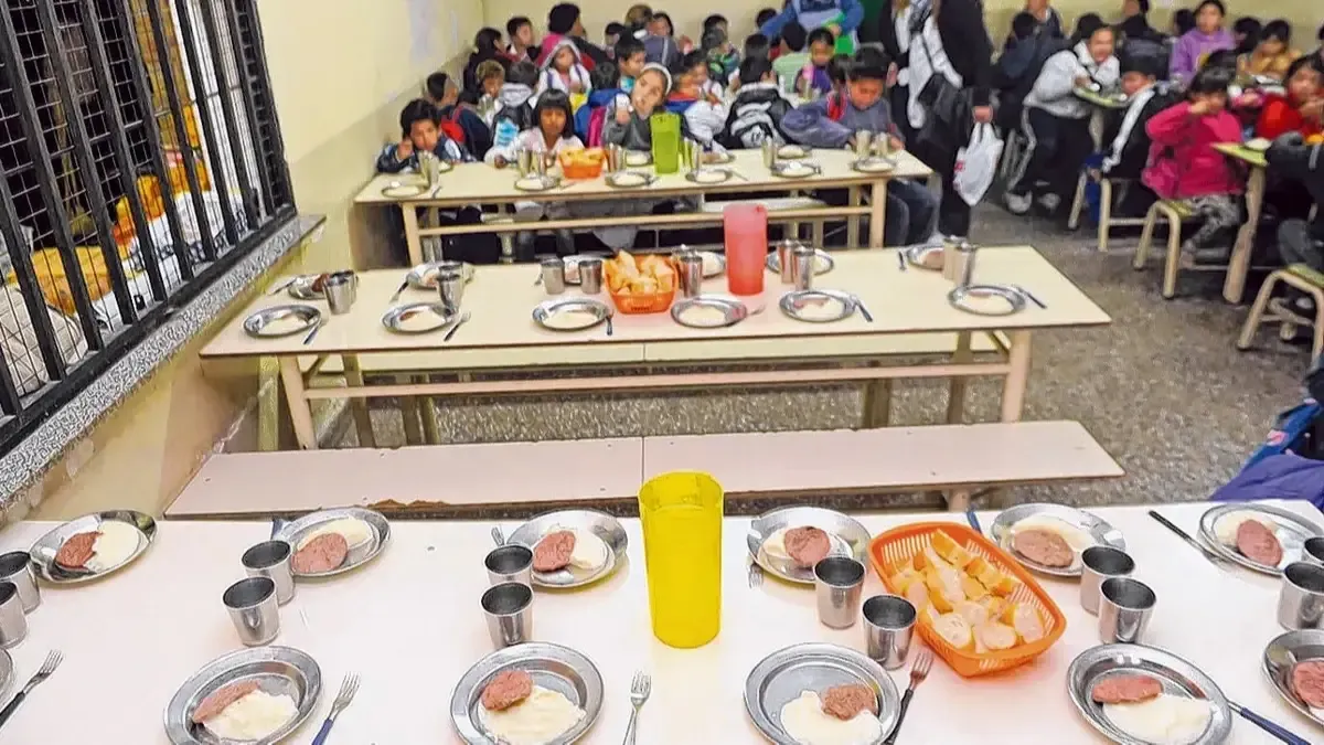Chicos sentados en mesas largas de un comedor escolar esperan la comida servida en platos de metal.