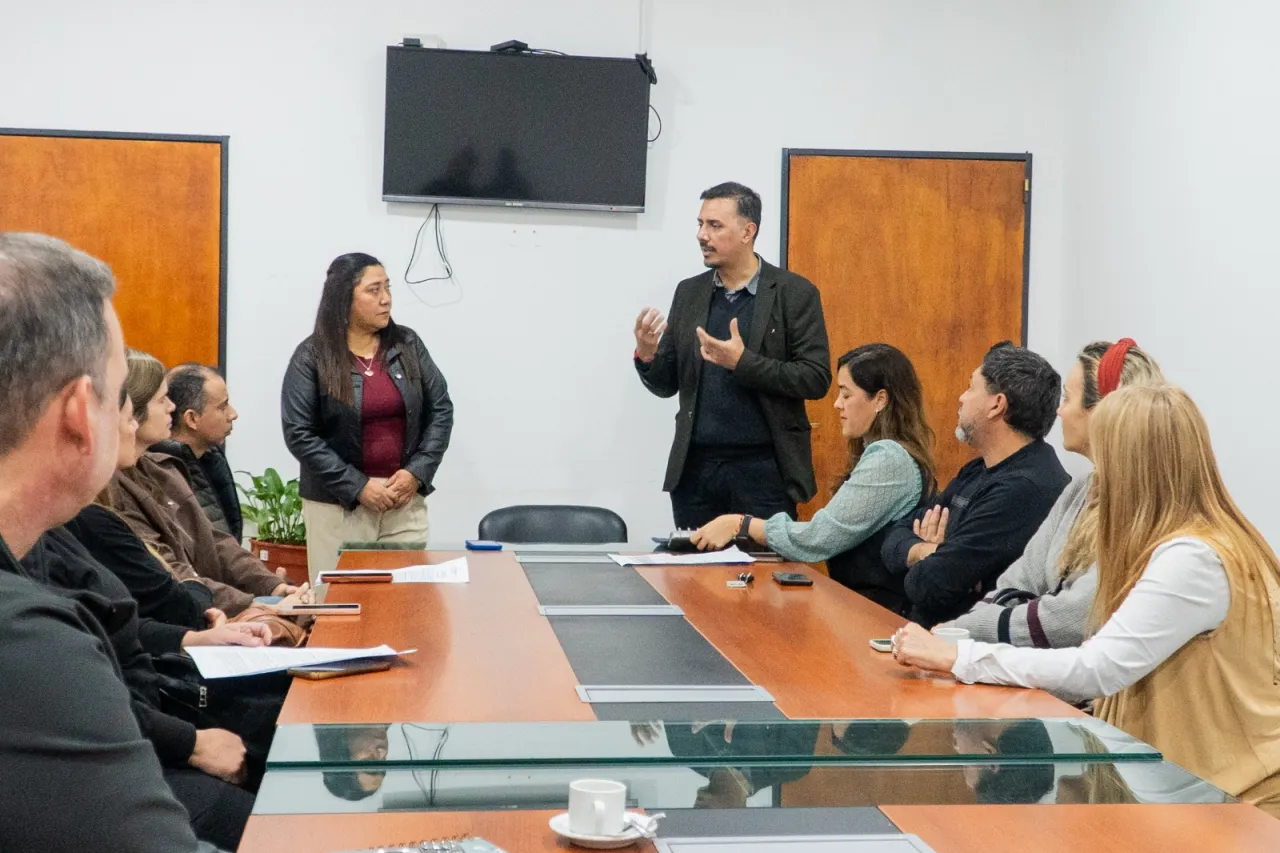 Un hombre de pie hablando con las manos ante un grupo de personas sentadas alrededor de una mesa de reuniones.