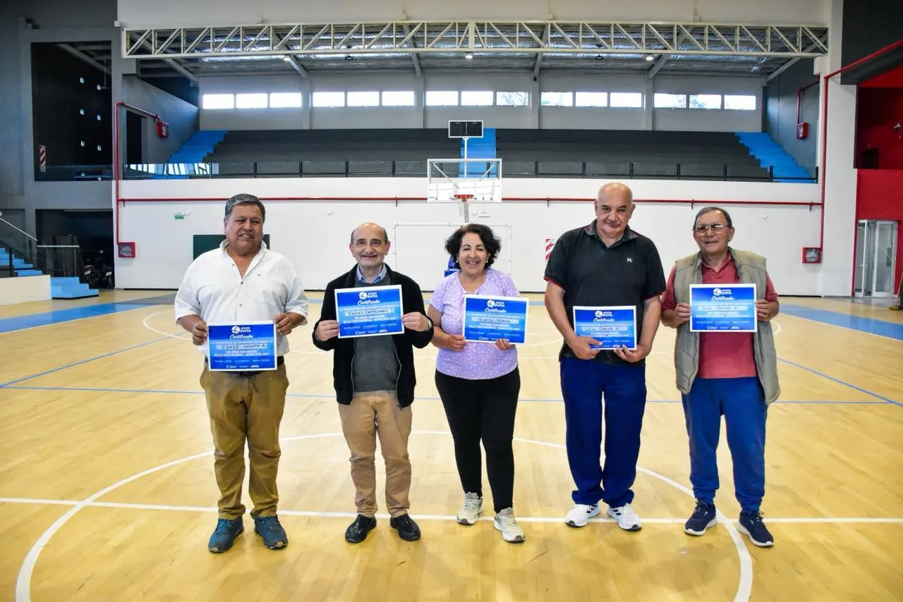 Cinco personas posan de pie en una cancha techada sosteniendo diplomas azules frente a la cámara.