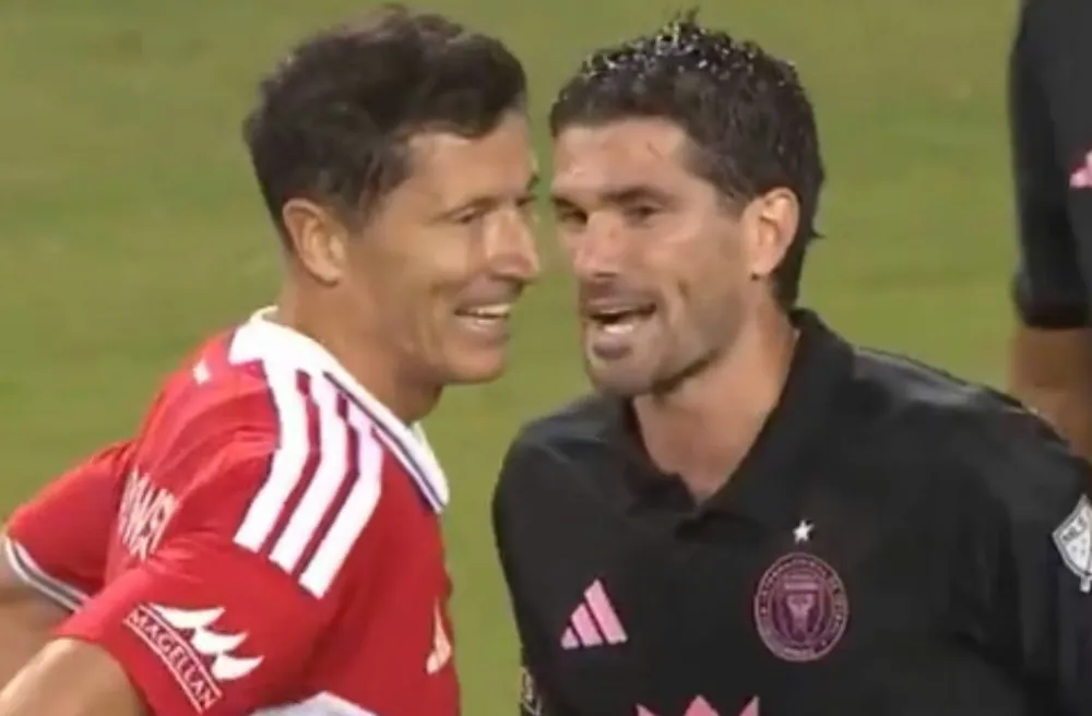 Dos futbolistas conversan de cerca en una cancha vistiendo camisetas deportivas.