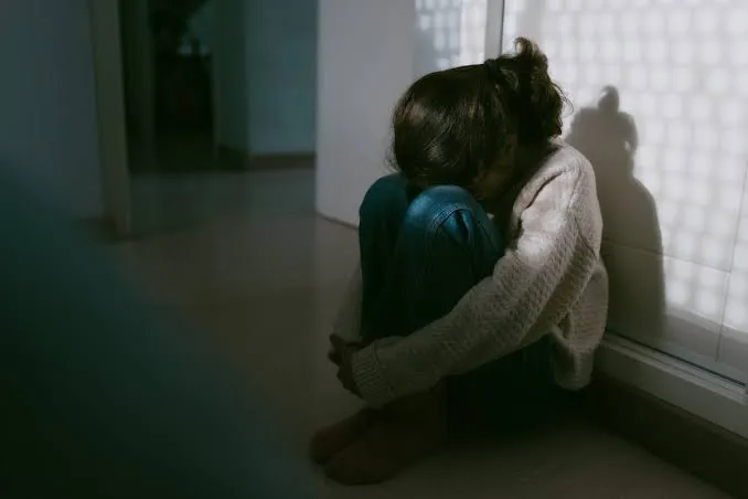 Niña sentada en el suelo abrazando sus rodillas en un rincón.