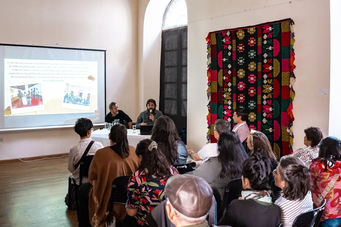 Un grupo de personas asiste a una disertación con proyector en una sala con un tapiz en la pared.