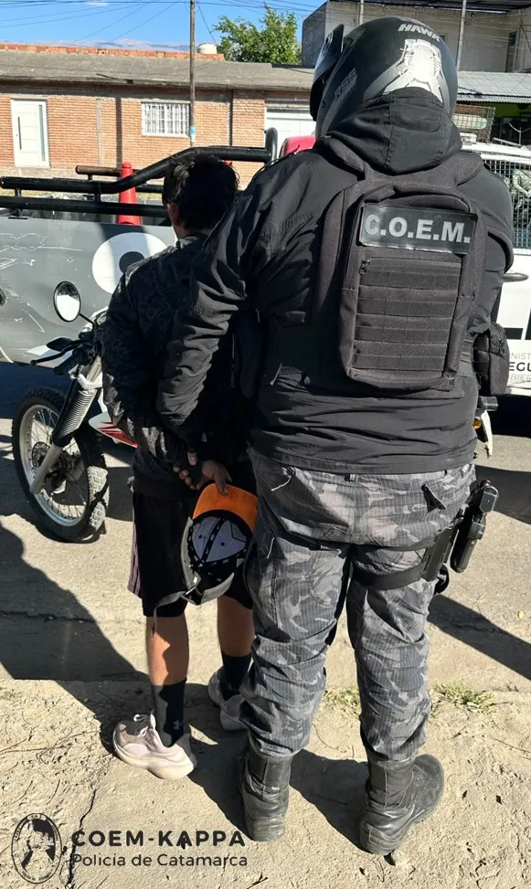 Un efectivo policial con chaleco táctico y casco sujeta a un joven de espaldas que lleva una gorra en la mano.