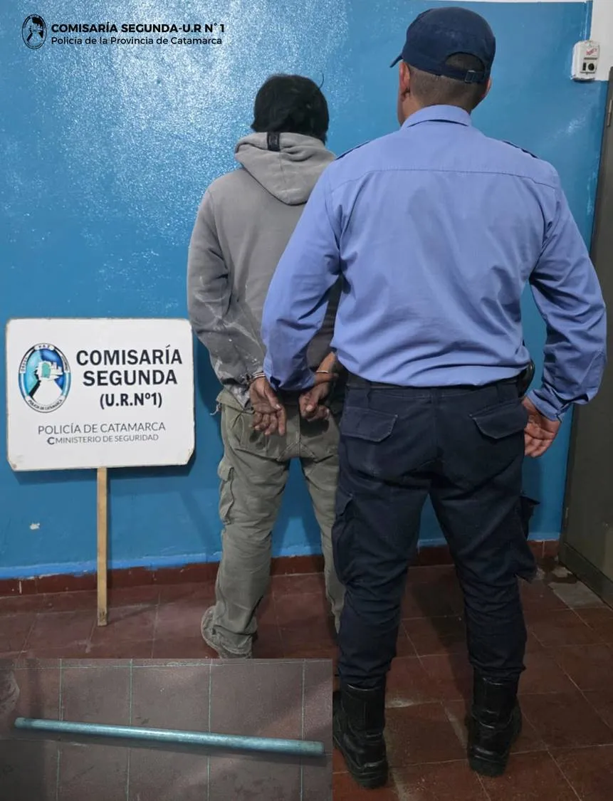Un policía de espaldas junto a un hombre esposado contra una pared azul y un caño en el suelo.