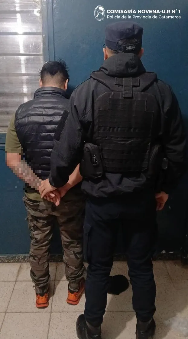 Un policia sostiene de las manos por la espalda a un hombre parado frente a una pared azul.