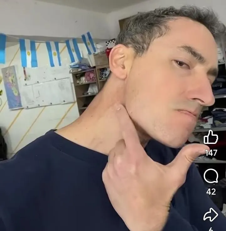 Un hombre en primer plano se señala con el dedo el lateral del cuello en una habitación con banderines argentinos.