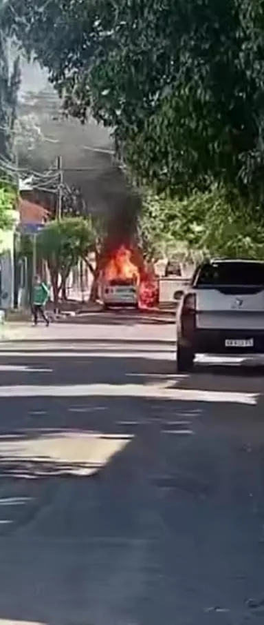 Un auto prendido fuego en una calle arbolada con humo negro, visto desde atrás de una camioneta estacionada.