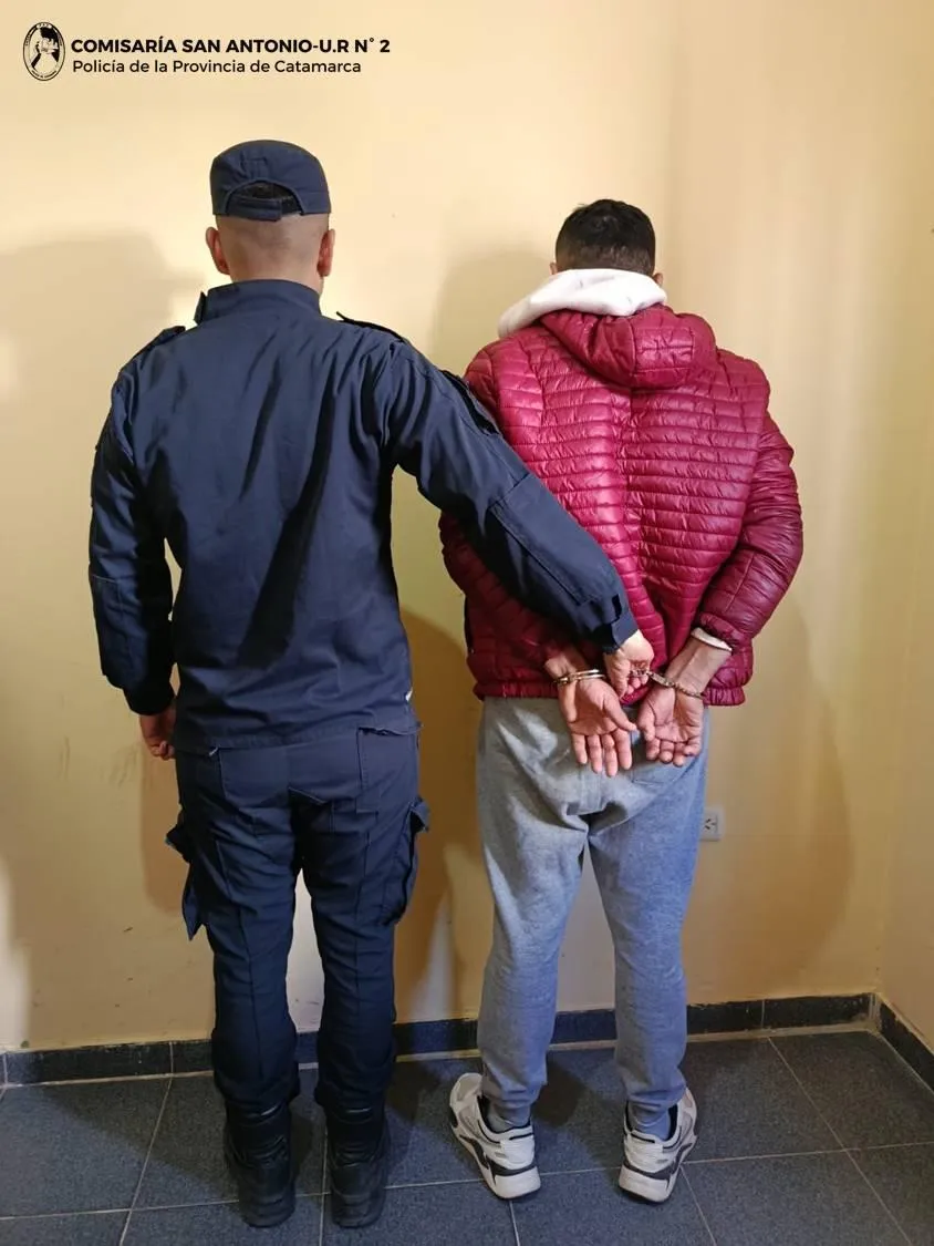Un oficial de policía sostiene de espaldas a un hombre esposado frente a una pared de interior.