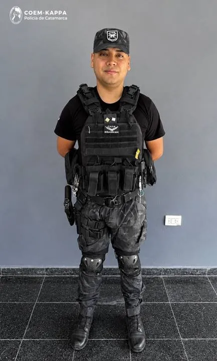 Un efectivo policial con uniforme táctico y chaleco antibalas posa de pie frente a una pared gris.