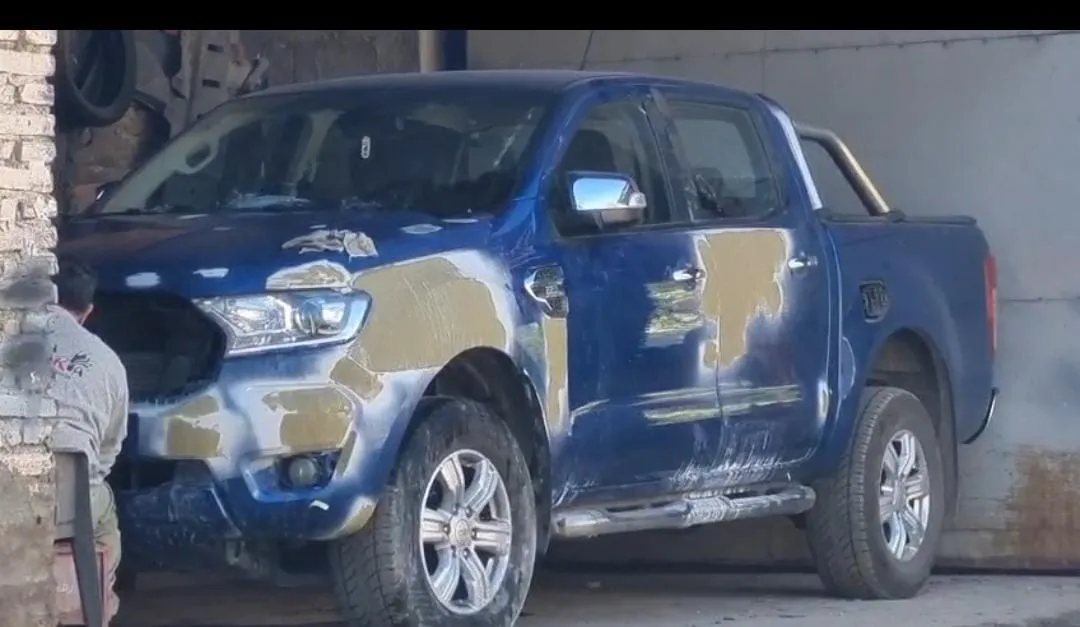 Un hombre sentado junto a una camioneta azul en proceso de chapa y pintura frente a un portón.