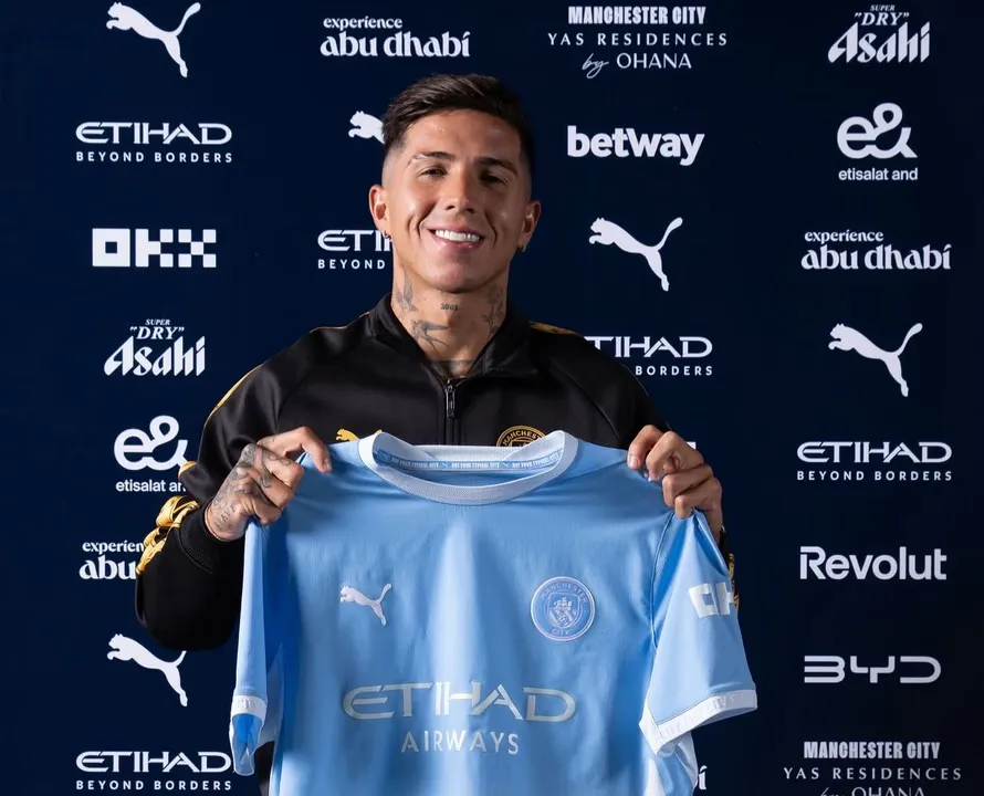 El futbolista Enzo Fernandez posa sonriente sosteniendo la camiseta celeste del Manchester City.