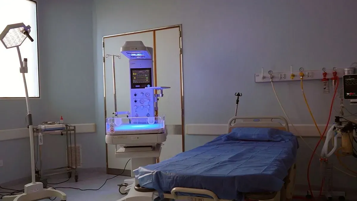 Una sala de hospital con una cama cubierta de azul, una cuna de terapia neonatal y equipamiento médico.