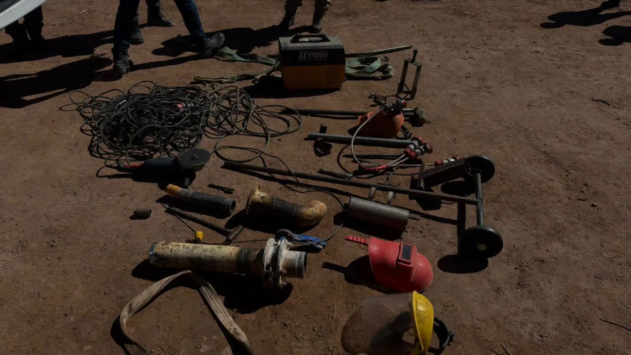 Distintas herramientas de soldadura, cables, amoladora y piezas metálicas yacen apoyadas sobre el suelo de tierra.