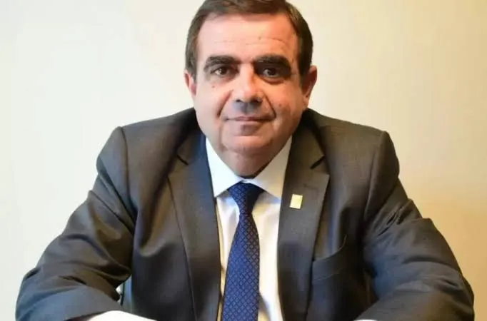 Hombre con traje gris, camisa blanca y corbata azul sentado frente a un escritorio.