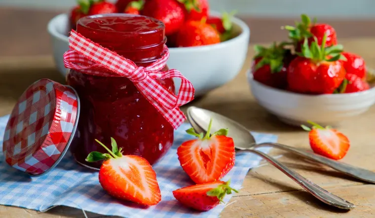 Un frasco de mermelada con cinta a cuadros se encuentra sobre un mantel junto a frutillas enteras y cortadas.