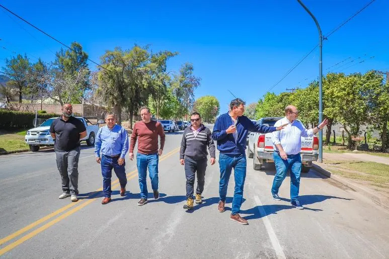 Un grupo de hombres camina por una calle asfaltada flanqueada por arboles y camionetas estacionadas.