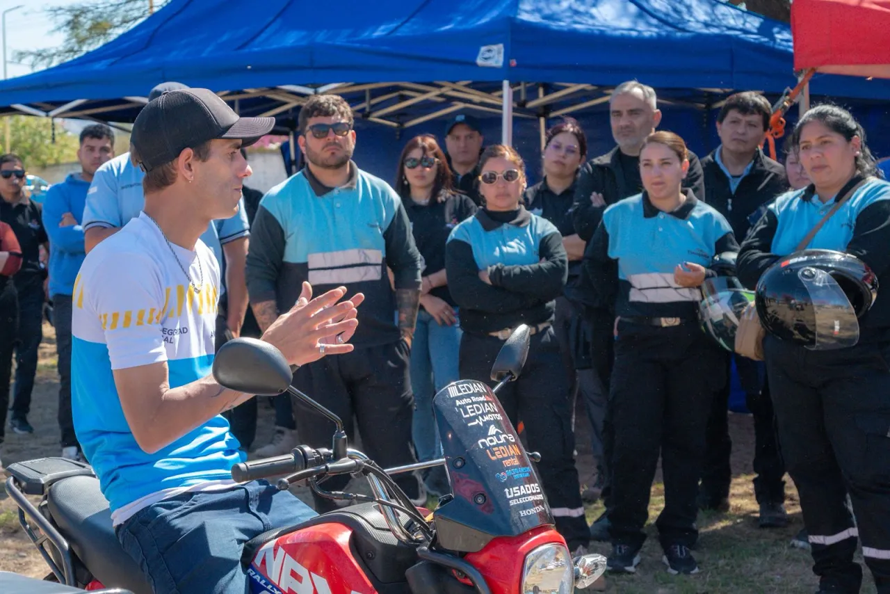 Un joven con gorra sobre una moto habla frente a un grupo de personas junto a un gazebo azul.