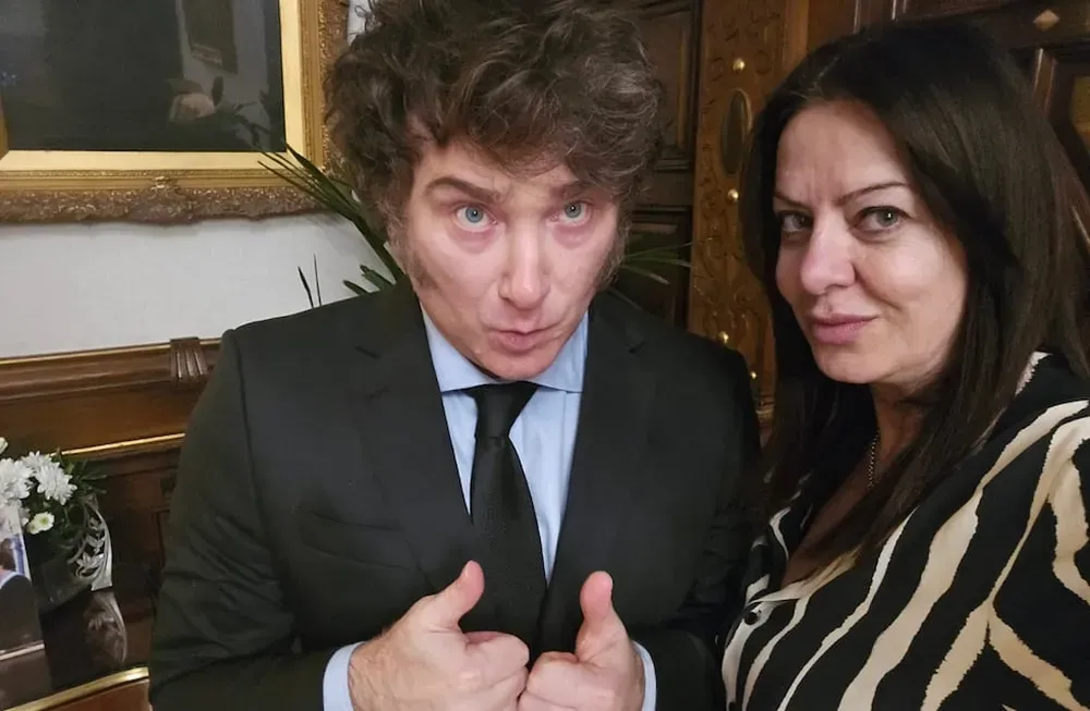 Javier Milei de traje hace el gesto de pulgares arriba junto a una mujer con blusa a rayas.