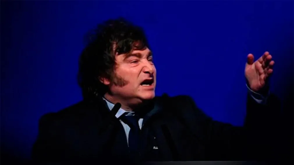 Javier Milei de traje y corbata gesticulando con la mano levantada frente a un fondo azul oscuro.