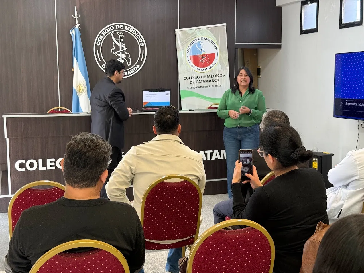 Una mujer expone de pie frente a un auditorio en una sala con banderas y un cartel del Colegio de Médicos.