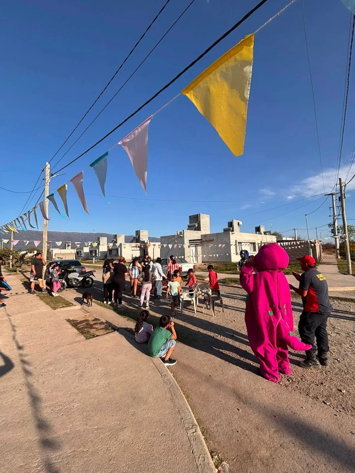 Personas y chicos se reúnen en una calle bajo guirnaldas junto a un muñeco inflable violeta y un bombero.