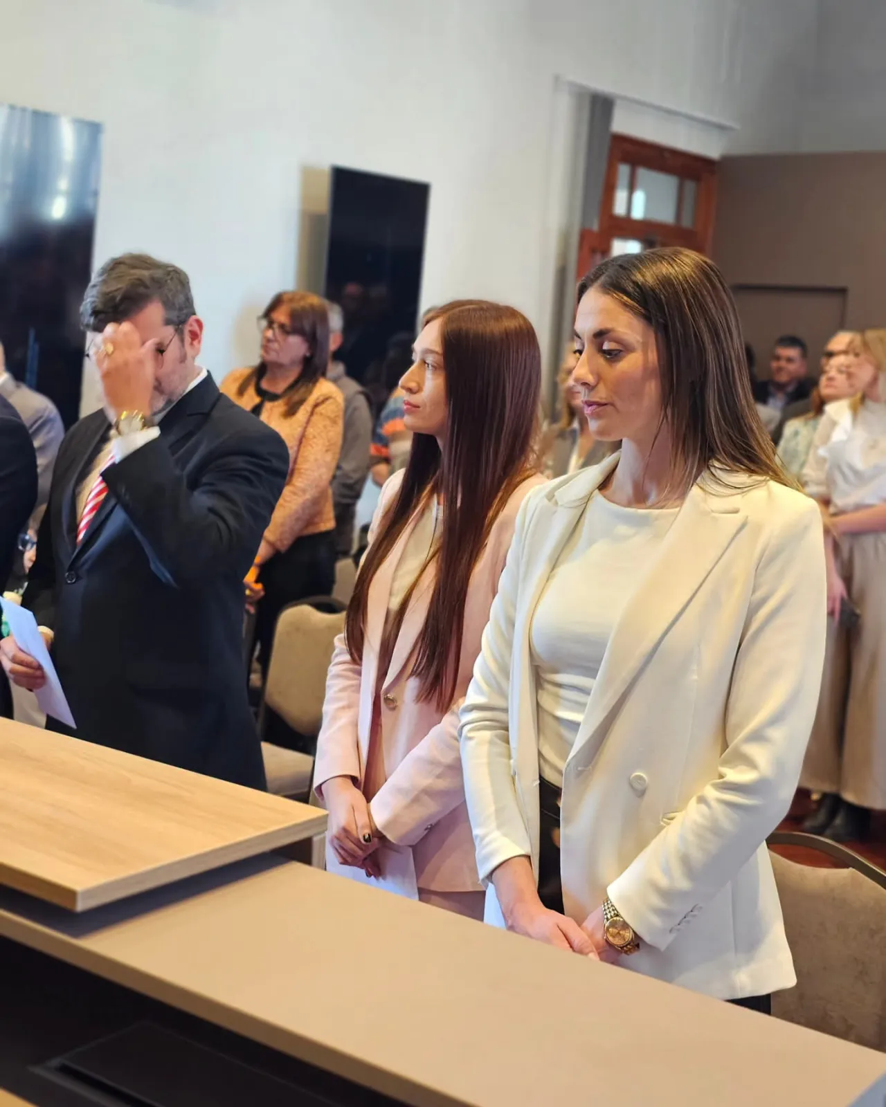 Dos mujeres vestidas formalmente de claro y un hombre de traje parados junto a un mostrador en un acto.