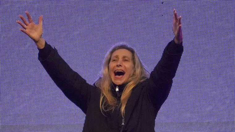 Karina Milei sonríe y levanta ambos brazos frente a una pantalla led de fondo violeta.