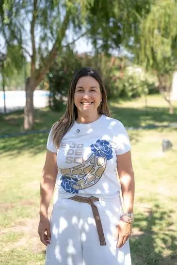 Una mujer sonriente parada al aire libre sobre el pasto, vestida con remera estampada y pantalón blanco.