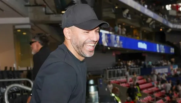 Sergio "Kun" Agüero sonríe usando una gorra negra en una tribuna deportiva.