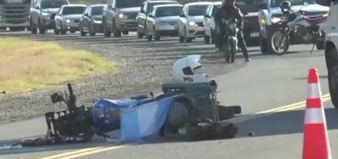 Una moto tirada sobre el asfalto junto a un casco, con autos frenados en fila de fondo y un cono en primer plano.