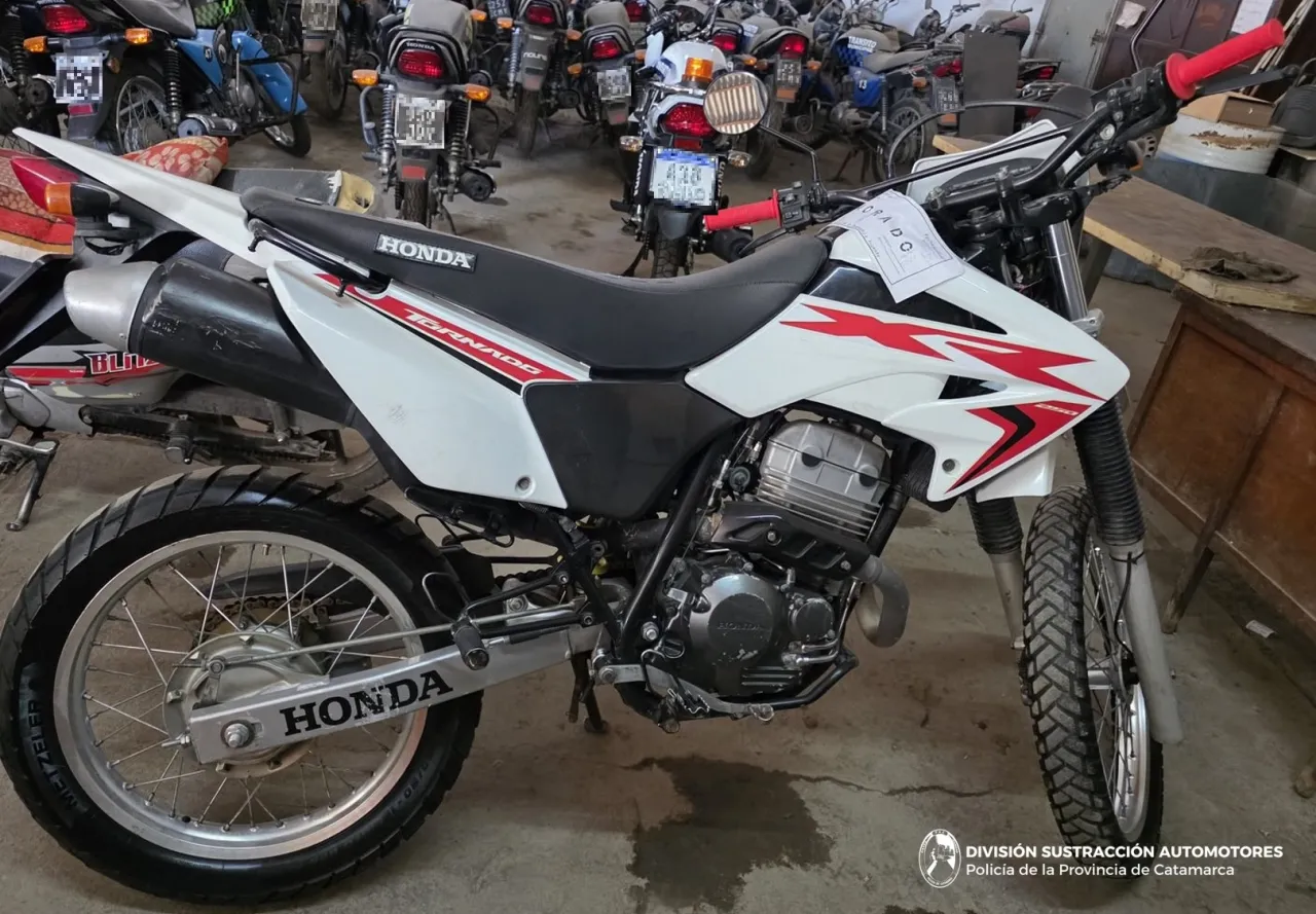 Una moto blanca marca Honda estacionada de costado adentro de un galpon junto a otros rodados.