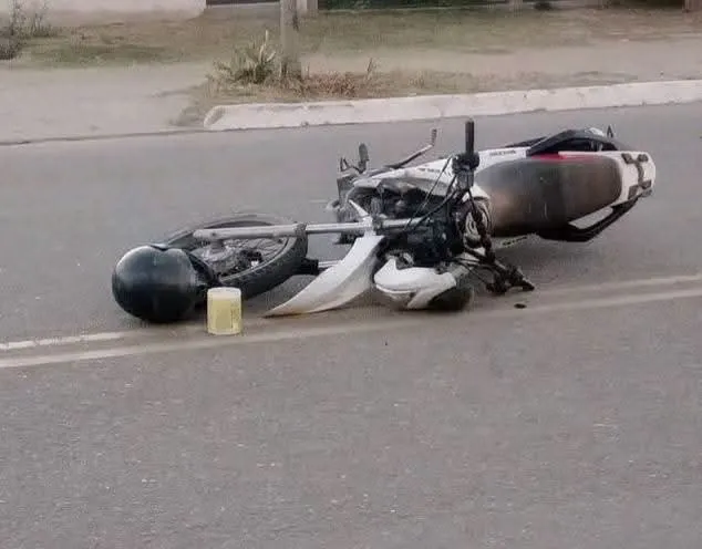 Una moto caída de costado sobre el asfalto junto a un casco negro y un recipiente plástico.
