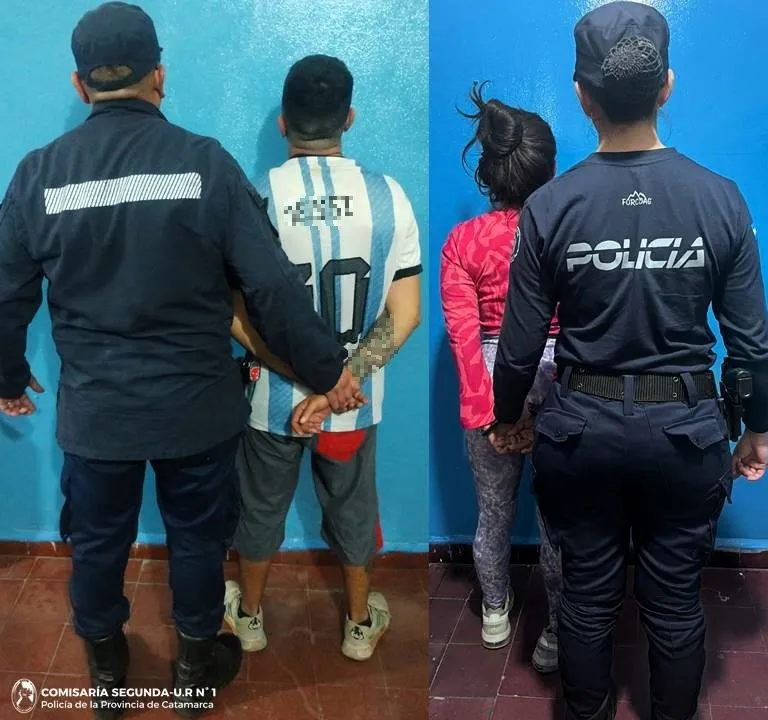 Dos policías de espaldas custodian a un hombre y una mujer demorados contra una pared azul.