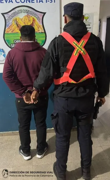 Un policía de espaldas custodia a un hombre esposado frente a un cartel de la Policía de Catamarca.