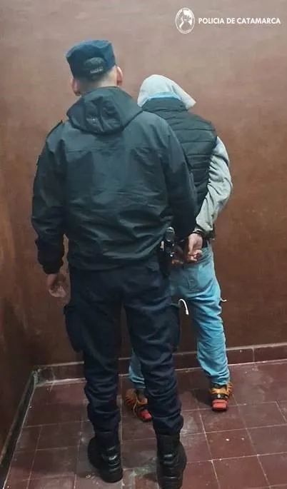 Un policia de espaldas sostiene a un hombre esposado contra una pared.