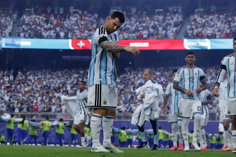 Lionel Messi acomodándose la manga con la camiseta de la selección argentina en una cancha de fútbol llena de gente.