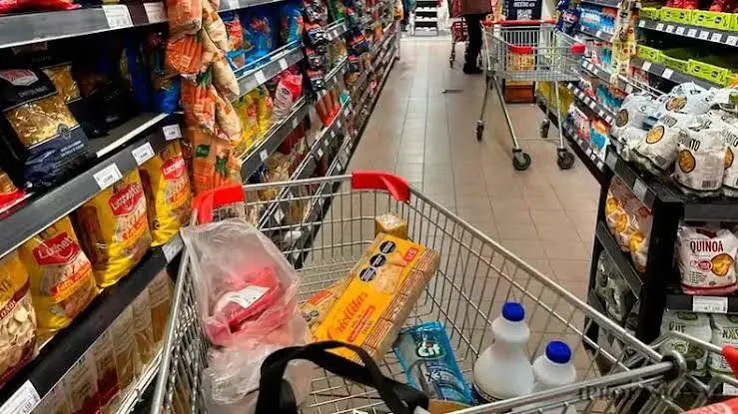 Un changuito de supermercado con mercadería en un pasillo entre góndolas llenas de paquetes de alimentos.