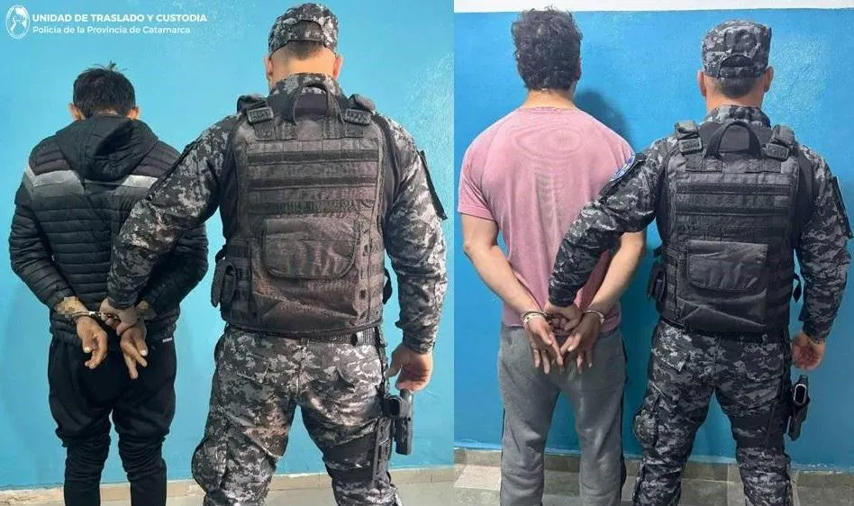 Dos oficiales uniformados de espaldas custodian a dos personas esposadas paradas frente a una pared celeste.