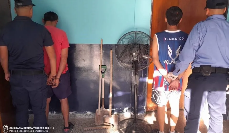 Dos policias custodian a dos hombres esposados de espaldas junto a un ventilador de pie y palas.