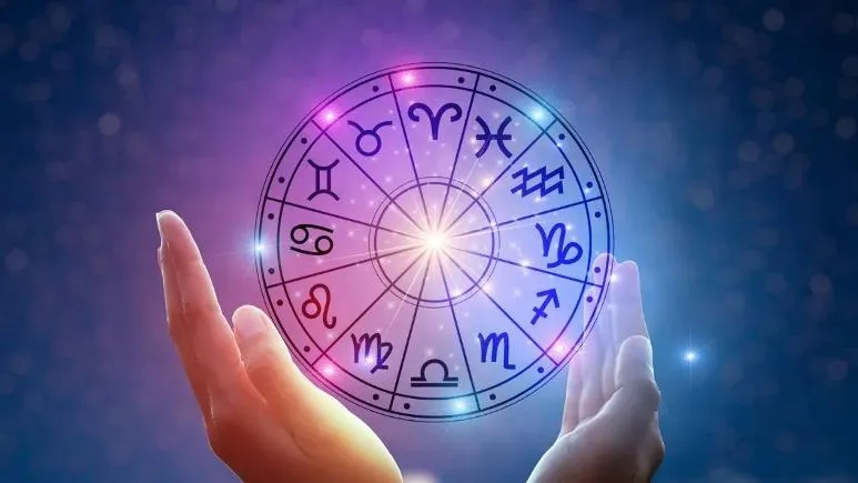 Dos manos sostienen una representacion circular iluminada con los doce signos del zodiaco en un fondo estrellado.