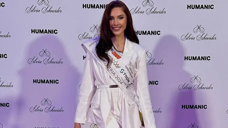 Lucia Sisic posa sonriendo con la banda de Miss Austria 2024 delante de un panel publicitario.