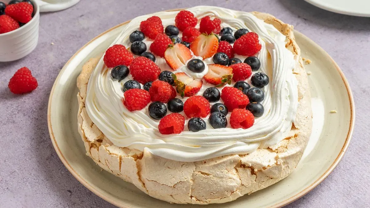 Una pavlova cubierta con crema chantilly, frutillas cortadas, arándanos y frambuesas sobre un plato claro.
