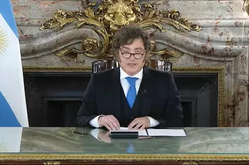 Javier Milei con traje y corbata azul habla sentado frente a un escritorio junto a la bandera argentina.