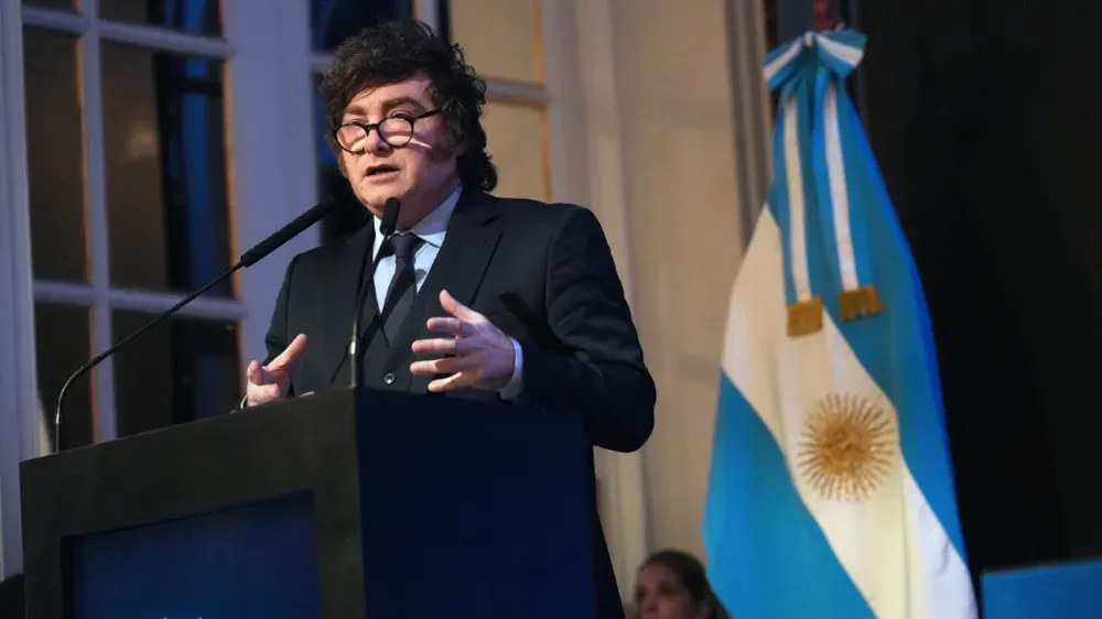 Javier Milei hablando desde un atril con microfonos junto a la bandera argentina.